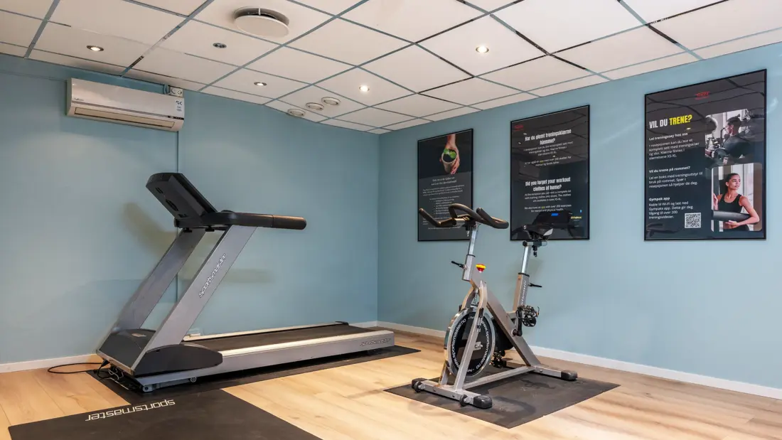 Treningsrom med tredemølle, trenings sykkel, stor trenings matte på gulvet. Blå vegger og tre plakater på veggen.