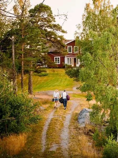 Rødt hus hytte ved kysten, to mennesker går på grusvei.
