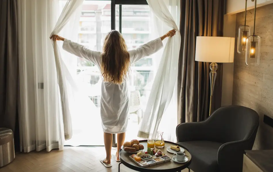 Dame som drar for gardiner i morgenkåpe mens frokosten står klar på lite rundt bord 