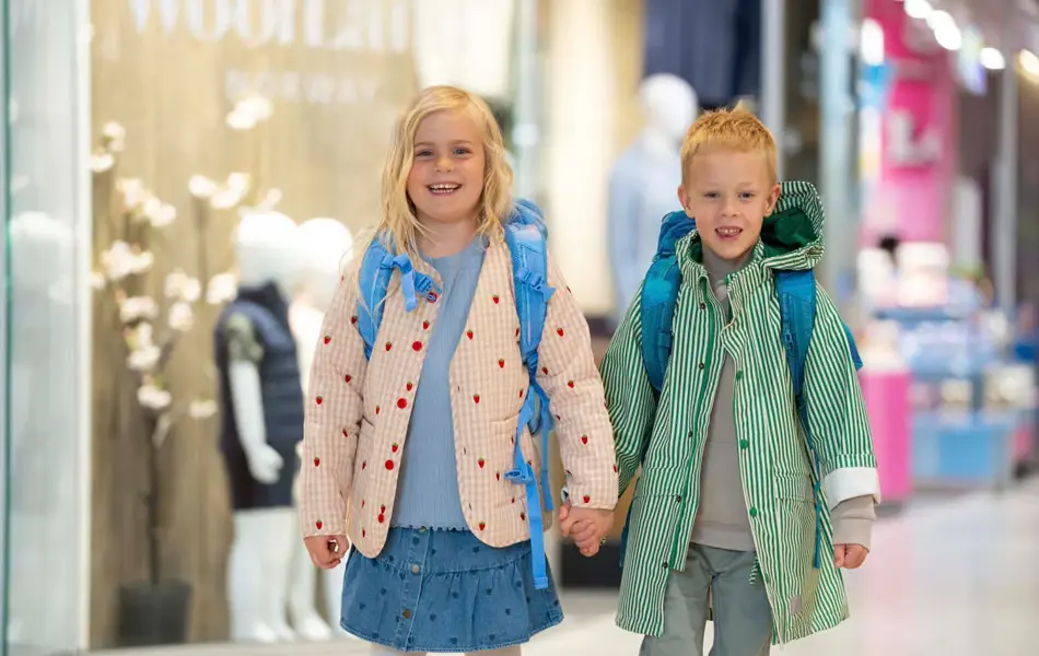 En jente og gutt holder hender på et kjøpesenter og har på yttertøy og skolesekk