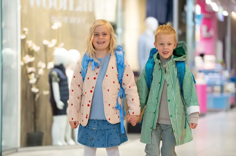 En jente og gutt holder hender på et kjøpesenter og har på yttertøy og skolesekk