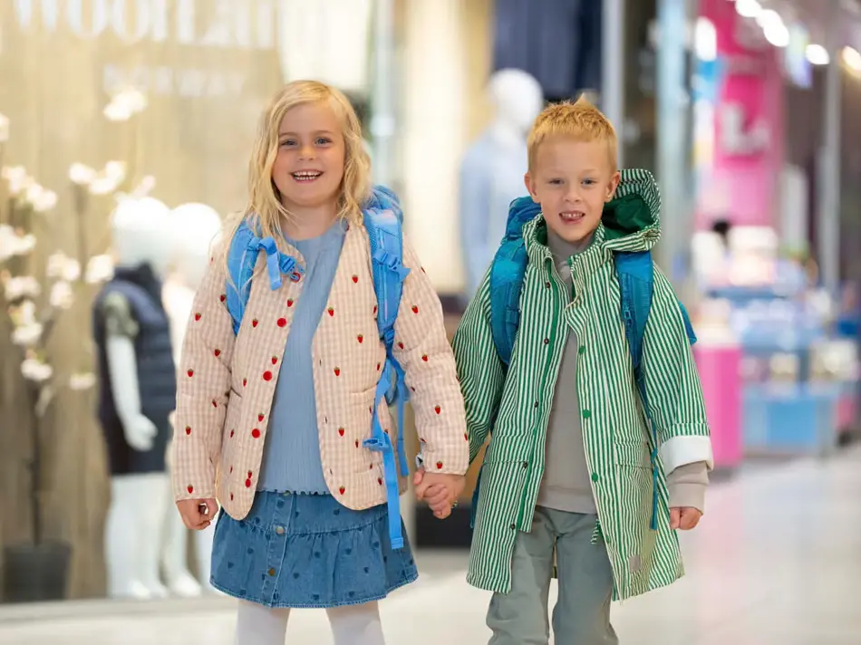 En jente og gutt holder hender på et kjøpesenter og har på yttertøy og skolesekk