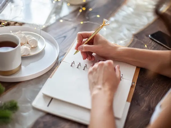 Young woman drinking tea and writing plans or goals for New Year 2021 while her daughter crafting Christmas trees from paper cone, yarns and buttons with placed stars and fairy lights on wooden table.
