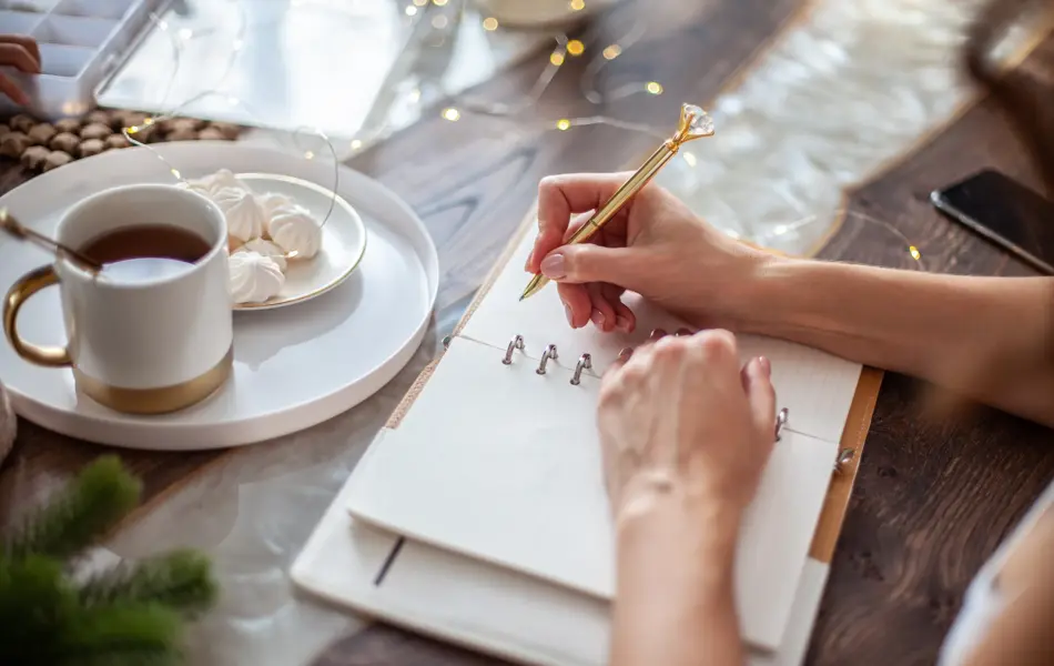 Young woman drinking tea and writing plans or goals for New Year 2021 while her daughter crafting Christmas trees from paper cone, yarns and buttons with placed stars and fairy lights on wooden table.