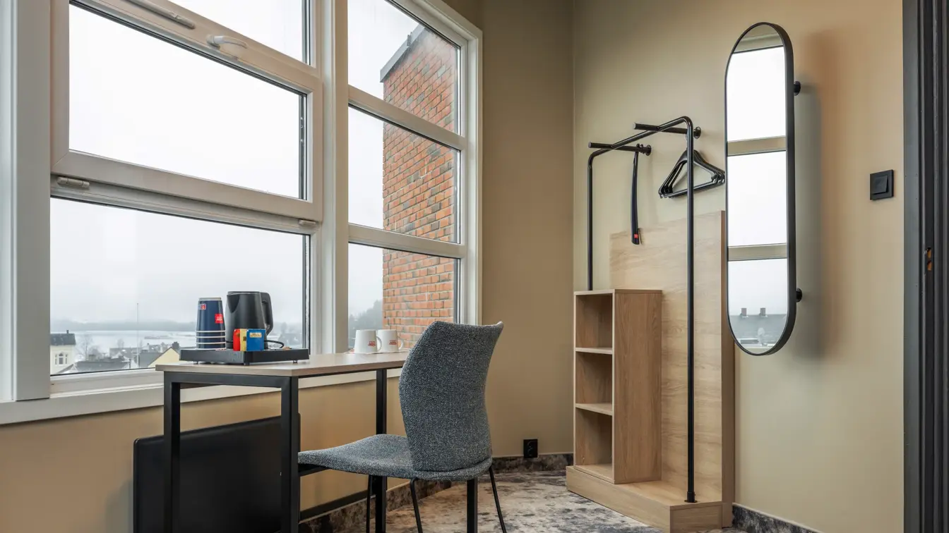 Hjørne av en junior suite, med skrivebord, stol, speil, åpen garderobe, stort vindu og teppegulv.