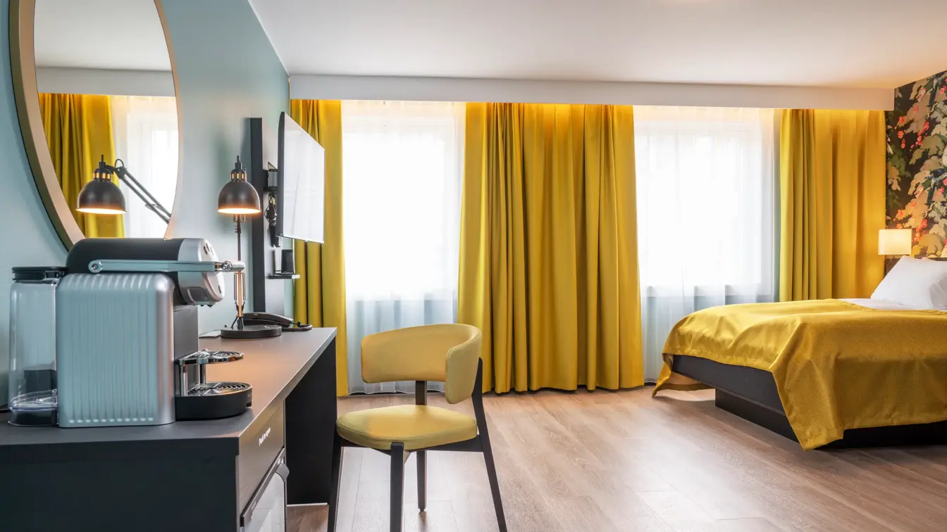 Skrivebord med kaffemaskin, leselampe og gul stol. TV, store vinduer med gule gardiner og seng med gult sengeteppe i business rom på Thon Hotel Bristol Bergen.