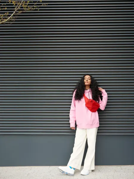 Dame med hvit bukse, rosa genser og rød veske poserer foran vegg 