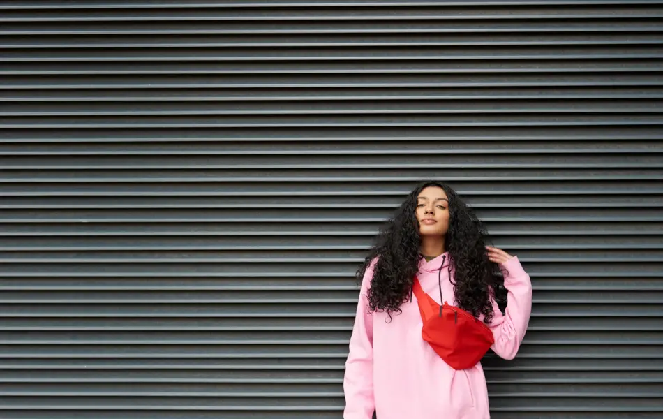 Dame med hvit bukse, rosa genser og rød veske poserer foran vegg 