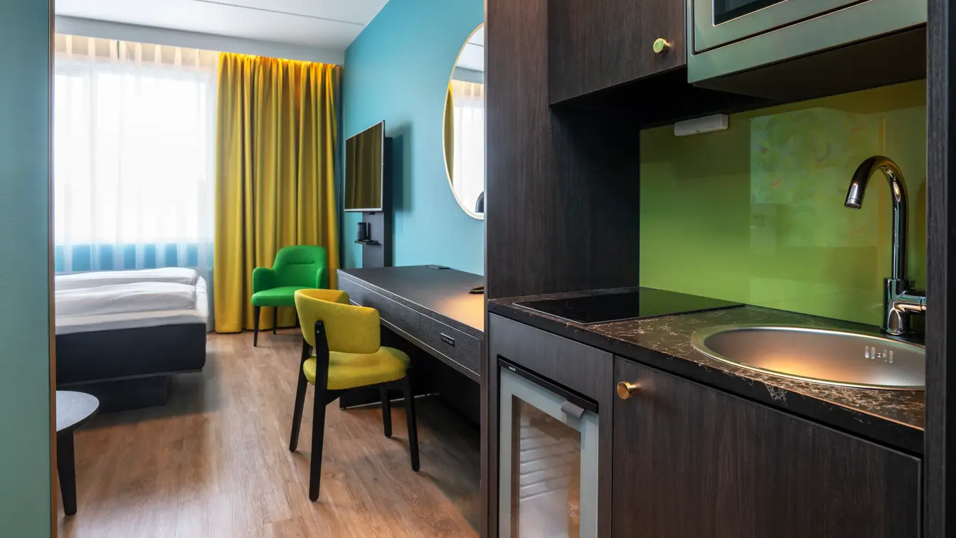 Dobbelseng, grønn og gul stol, gule gardiner, vindu, blå vegg, speil, kjøkken, skrivebord