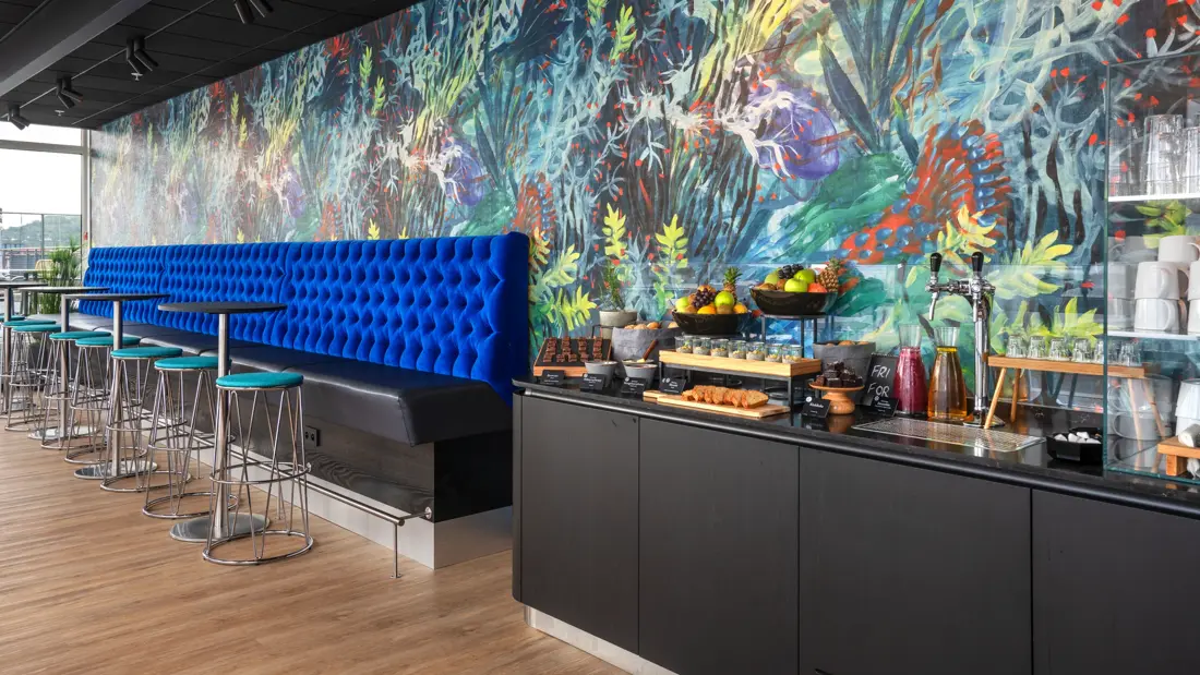 Fargerik tapetvegg, parkettgulv, en blå sofa, et svart rundt bord med turkise barkrakker, bordbenk med div mat.