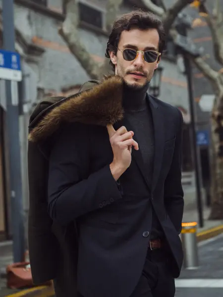 Halvportrett av mann med sort dress og solbriller holder jakke med pelskrage over skulderen ute på gata 