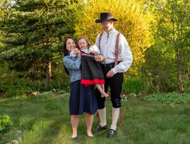 Mor i kjole og far i bunad holder lite barn med festdrakt. Står i hage og smiler mot kamera. Foto til artikkel om tips til antrekk 17. mai.