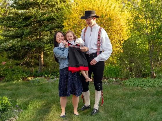 Mor i kjole og far i bunad holder lite barn med festdrakt. Står i hage og smiler mot kamera. Foto til artikkel om tips til antrekk 17. mai.