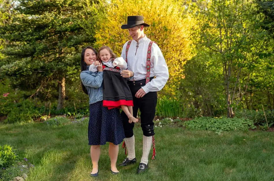 Mor i kjole og far i bunad holder lite barn med festdrakt. Står i hage og smiler mot kamera. Foto til artikkel om tips til antrekk 17. mai.