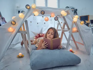 Little 10 year old girl reading classic book under her home-made tent inside the living room.