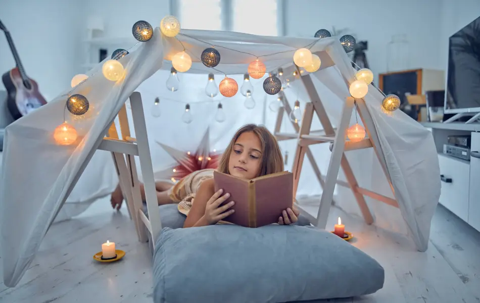 Little 10 year old girl reading classic book under her home-made tent inside the living room.