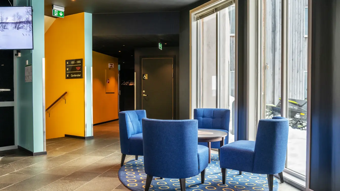 Lobbyområde med fire blå lenestoler rundt et rundt bord, rundt blått teppe under, og store vinduer ut.