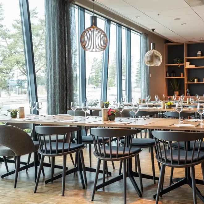 Langbord hos Restaurant Jaktlaget. Bordene er dekket med glass, servietter og bestikk, samt bordpynt. Restauranten får dagslys inn fra de store vindusflatene som går fra gulv til tak.