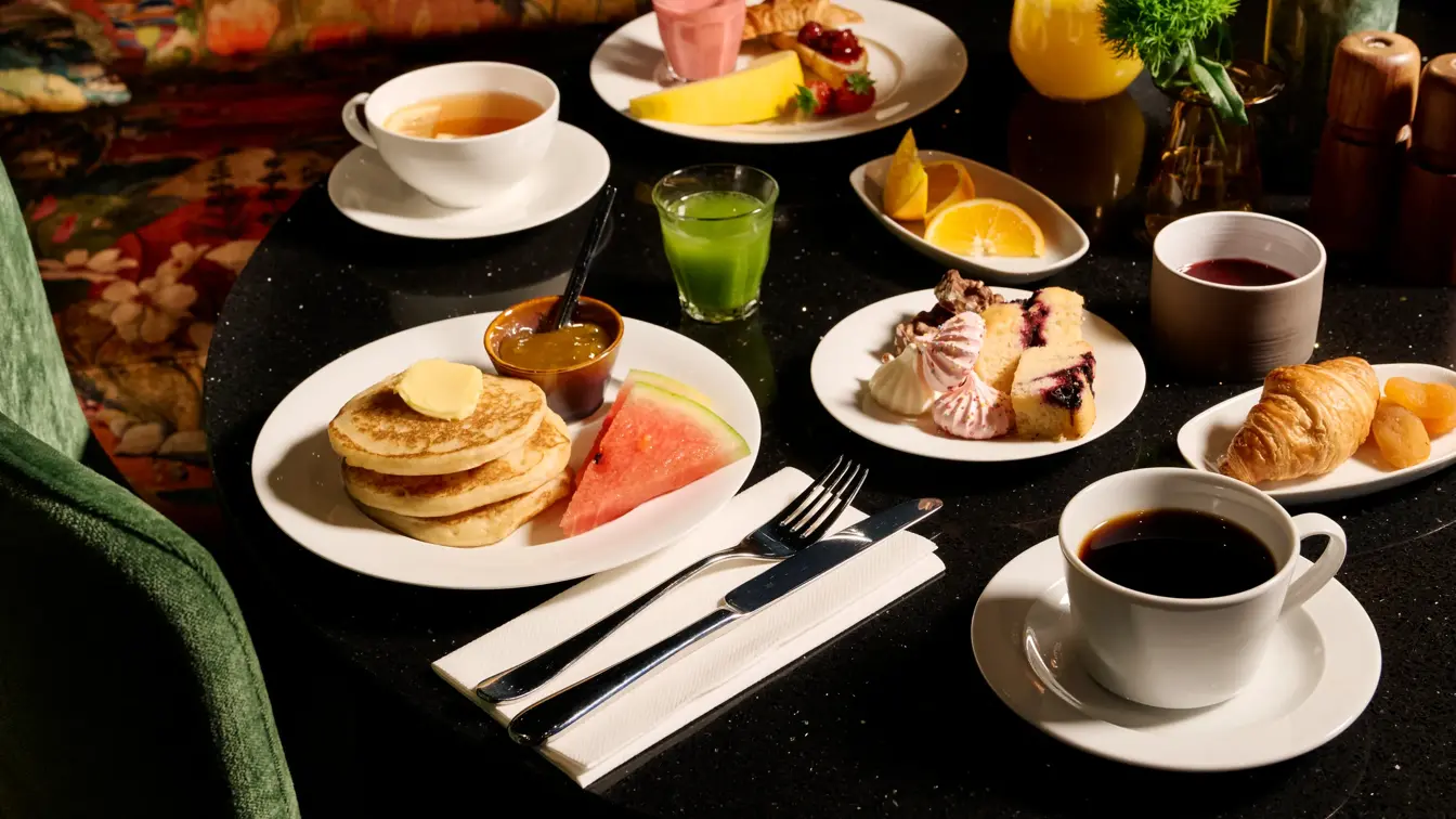 Et frokostbord med en tallerken med pannekaker, smør, marmelade og vannmelon. Kaffe, crossiant, te og grønn juice.