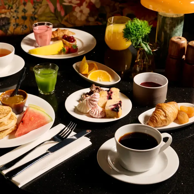Et frokostbord med en tallerken med pannekaker, smør, marmelade og vannmelon. Kaffe, crossiant, te og grønn juice.
