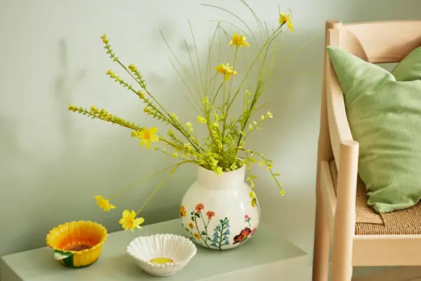 Lite bord med gul bolle, hvit bolle, blomstrete vase med gule blomster, stol med grønn pute