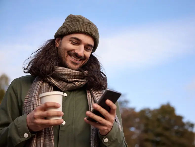 Mann med olivengrønn lue, olivengrønn jakke og beige rutete skjerf står utendørs og ser på mobilen og holder en kaffekopp mens han smiler.