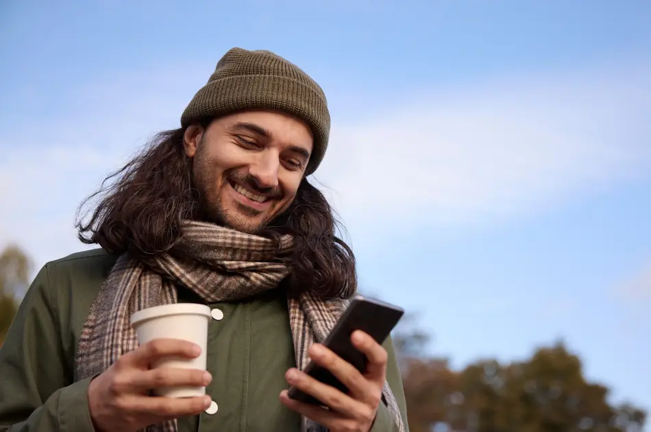 Mann med olivengrønn lue, olivengrønn jakke og beige rutete skjerf står utendørs og ser på mobilen og holder en kaffekopp mens han smiler.