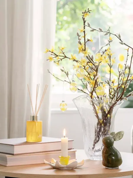 Bord med vase med grener med gule blomster, lysestake, bøker og duftpinner.