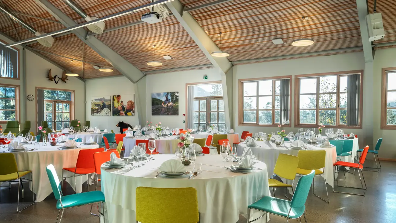 Selskapslokale dekket opp med runde bord, hvite duker, fargerike stoler, vinduer og høyt under taket.