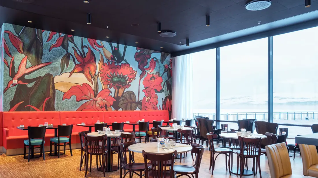 Knall rød sofa med røde store blomster på tapet i bagrunnen, sitteplasser med runde bord og utsikt mot havet gjennom store vinduer i restaurant på Thon Hotel Kirkenes ved kaia i Kirkenes sentrum