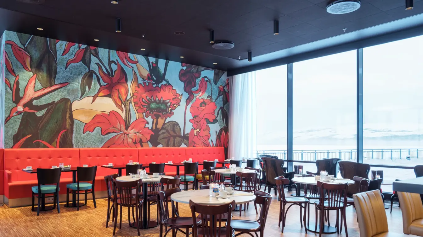 Knall rød sofa med røde store blomster på tapet i bagrunnen, sitteplasser med runde bord og utsikt mot havet gjennom store vinduer i restaurant på Thon Hotel Kirkenes ved kaia i Kirkenes sentrum