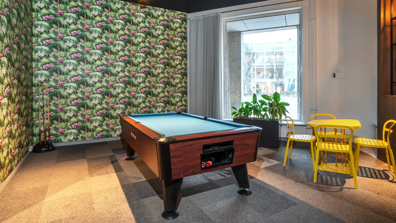 Biljardbord, tapetsert vegg med blomster, biljardkøer på stativ i hjørnet, 4 gule stoler rundt gult bord, stort vindu.