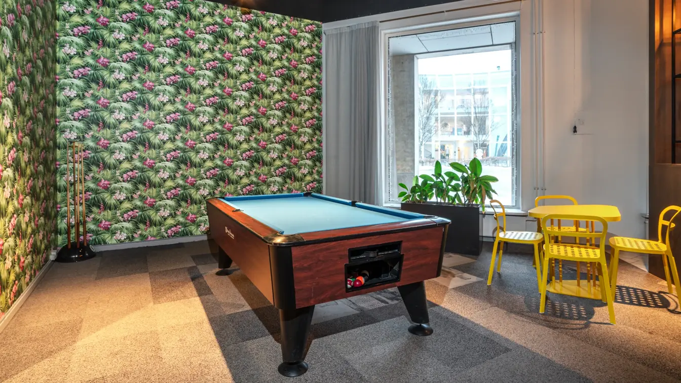 Biljardbord, tapetsert vegg med blomster, biljardkøer på stativ i hjørnet, 4 gule stoler rundt gult bord, stort vindu.