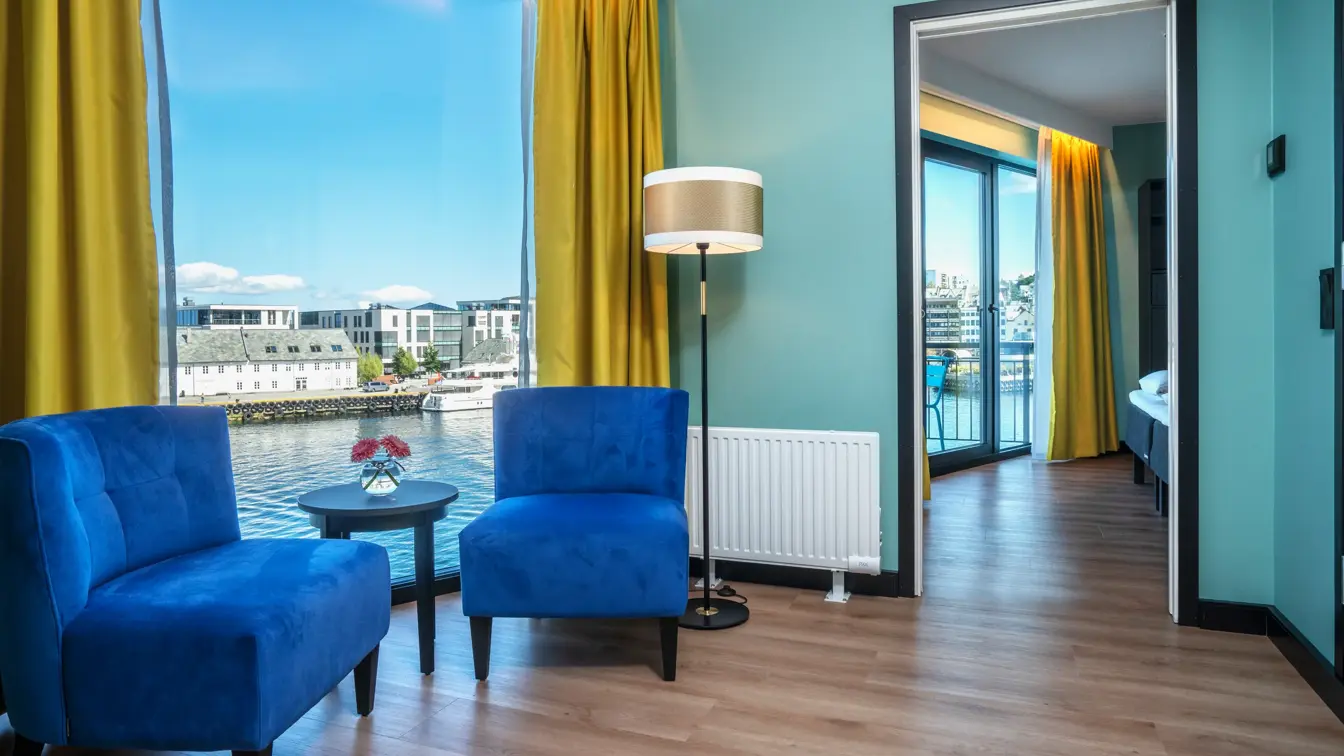 Gule gardiner, panoramavindu med utsikt over vannet, to blå lenestoler, salongbord, turkis vegg