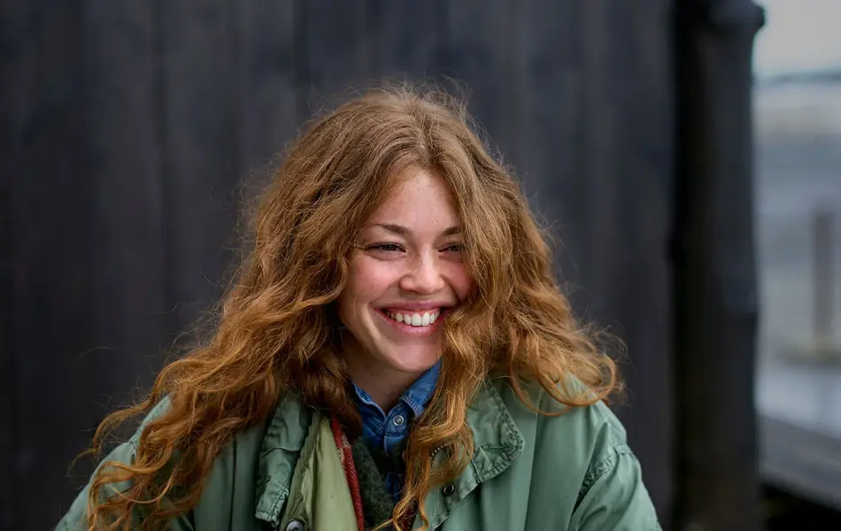 Dame i stor, grønn jakke og langt, bølgete hår smiler utendørs 