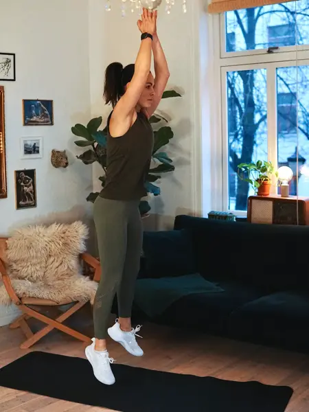 treningsekspert Anna Johansen trener på matte på gulvet, mørkegrønn sofa i bakgrunnen, lys stol med pels oppi, str grønn plante, bilder på veggen