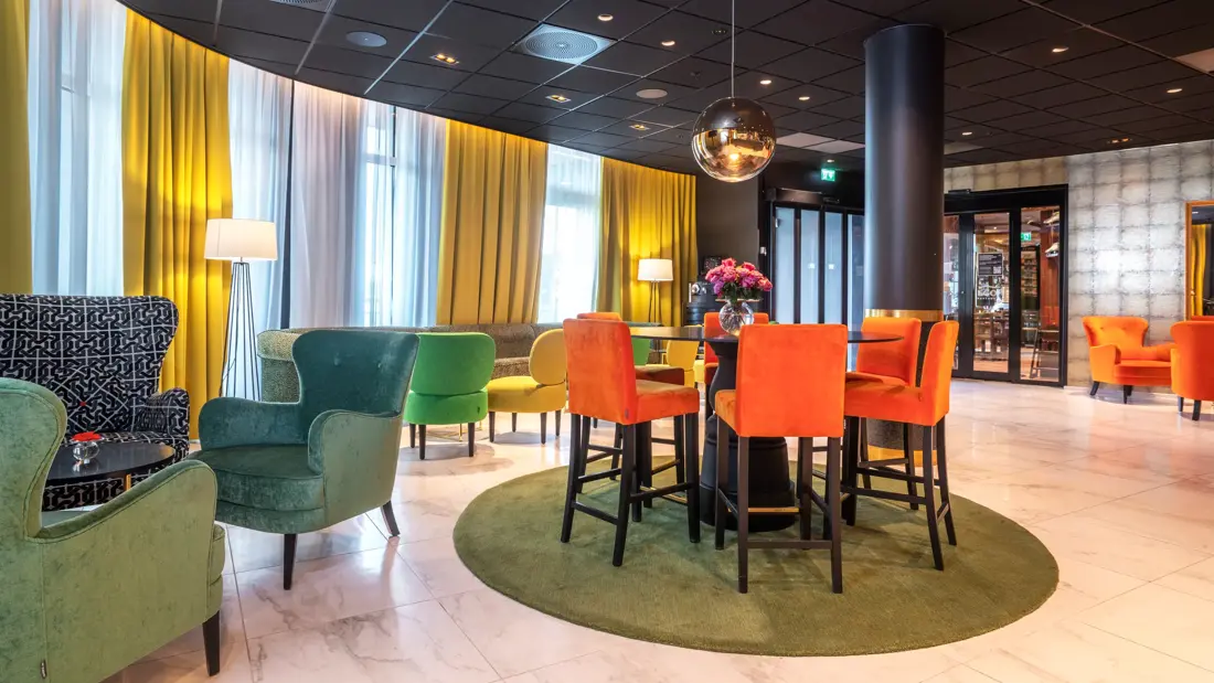 Sittelounge, oransje stoler, runde bord, grønne og blå stoler, vindu fra gulv til tak, gule gardiner, parkettgulv. 