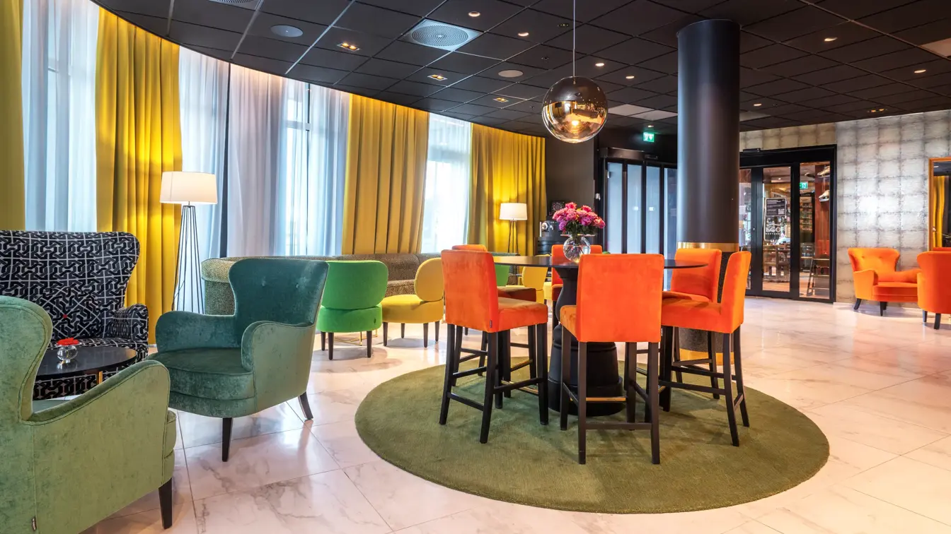 Sittelounge, oransje stoler, runde bord, grønne og blå stoler, vindu fra gulv til tak, gule gardiner, parkettgulv. 