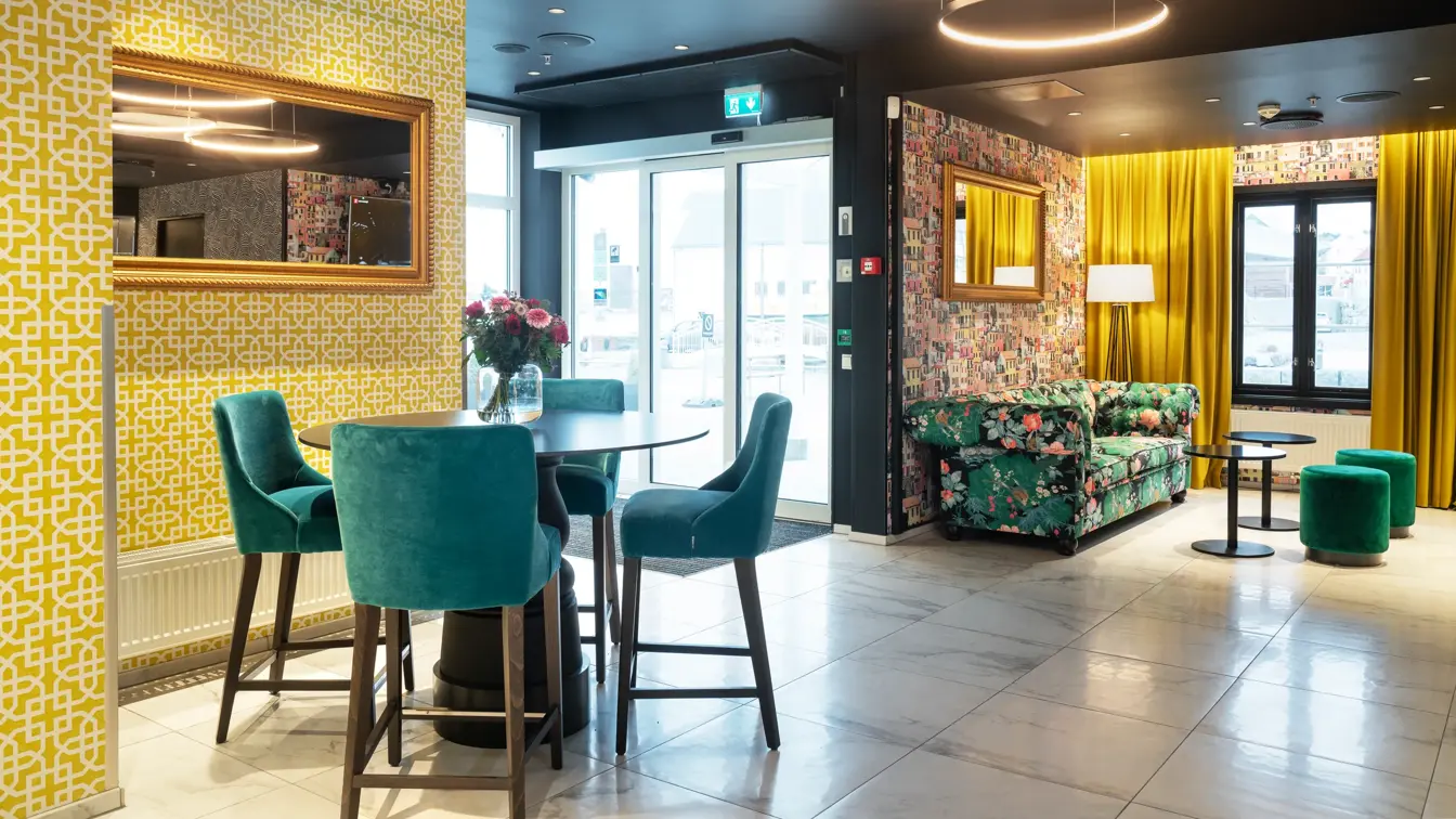 Gult tapetvegg med mønster, gule gardiner, fire turkise stoler, rundt svart bord, utgangsdør, sofa to grønne puffer.