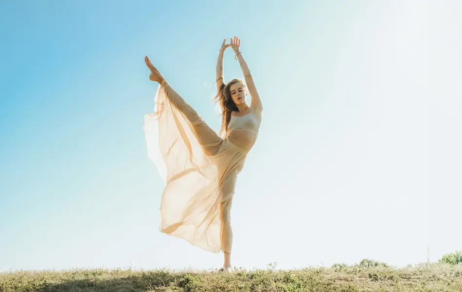 En kvinne danser utendørs i en gjennomsiktig drakt. Hun løfter det høyre benet høyt og holder begge armene i luften. 