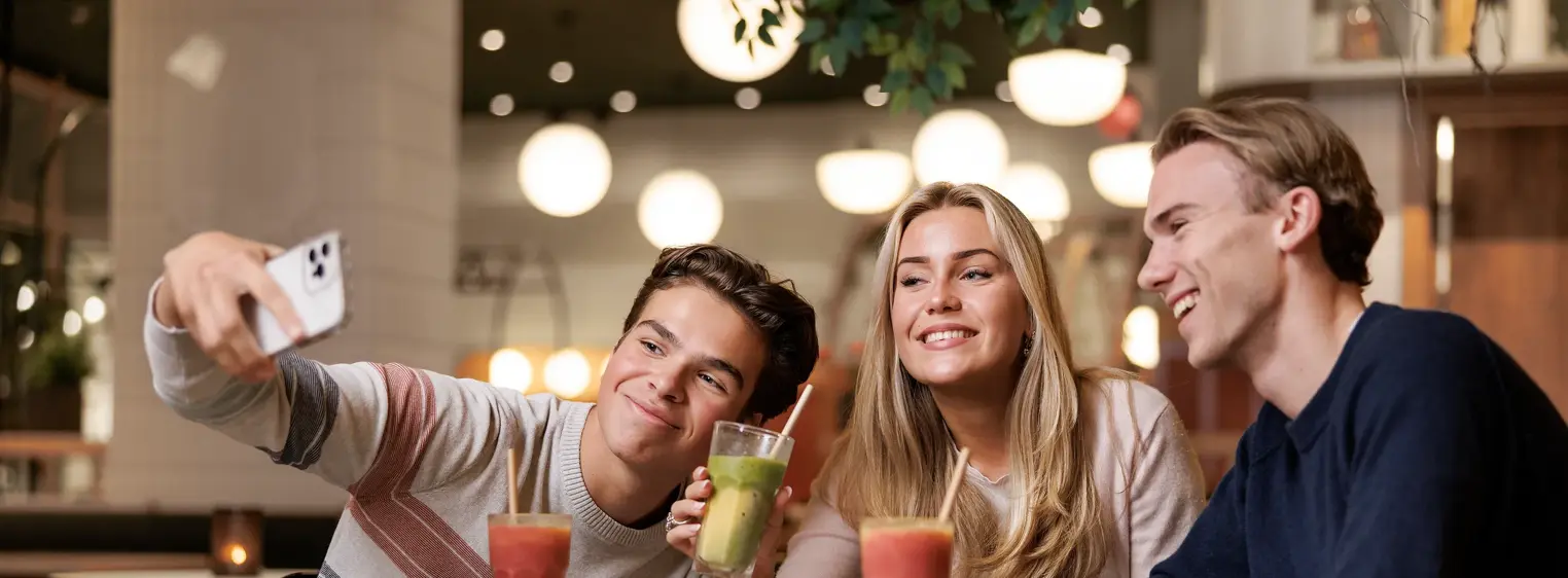 To gutter og en jente som sitter ved et bord i en kafé og tar bilder av seg selv med en mobil