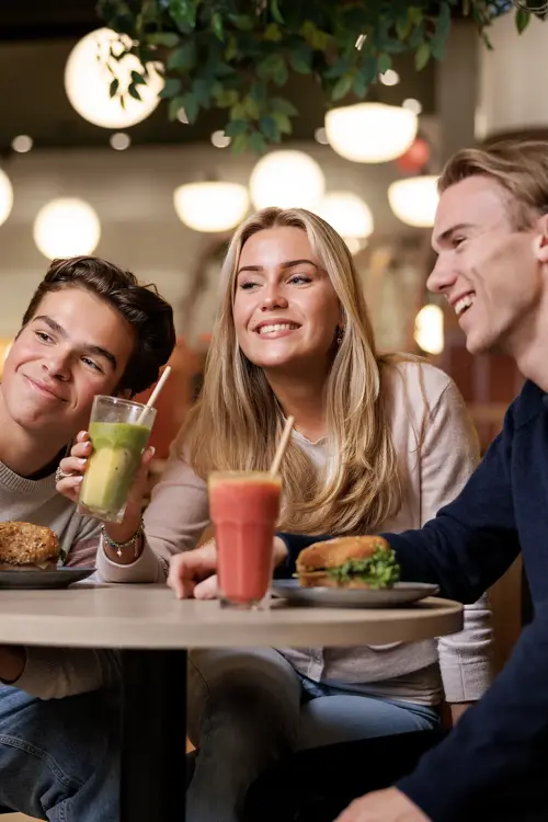 To gutter og en jente som sitter ved et bord i en kafé og tar bilder av seg selv med en mobil