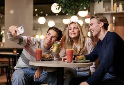 To gutter og en jente som sitter ved et bord i en kafé og tar bilder av seg selv med en mobil
