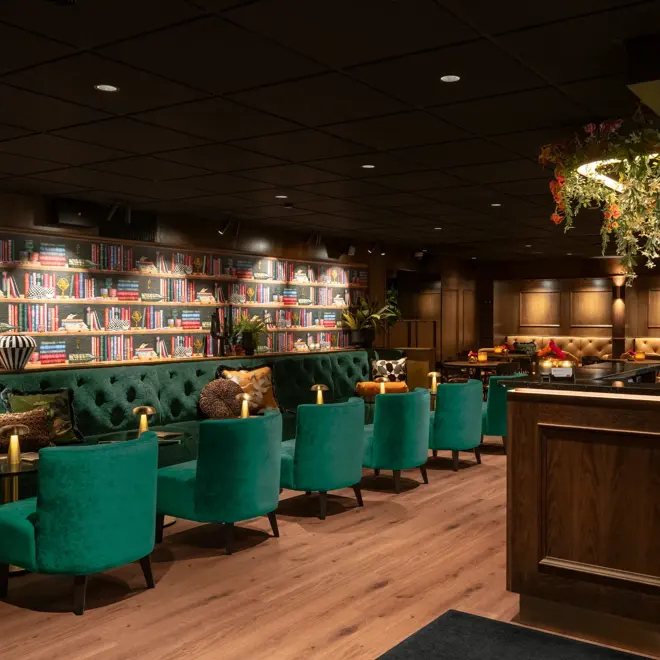 Lobby og barområde med bardisk i mørkt tre, lenestoler og sofa i dyp grønnfarge. Dempet belysning.