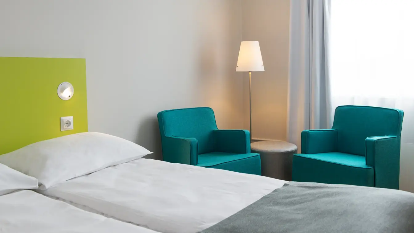 Nærbilde av Standard Room Twin på Thon Hotel Trondheim. Hvit vegg, limegrønt sengegavl, to oppredde enkeltsenger, to turkise lenestoler, lampe og et lite rundt bord.