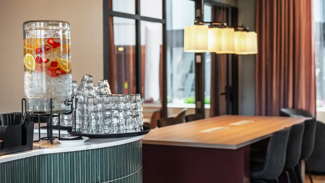 Vanndispenser og vannglass på resepsjonsdisk. Arbeidsbord med tilhørende stoler i bakgrunnen.