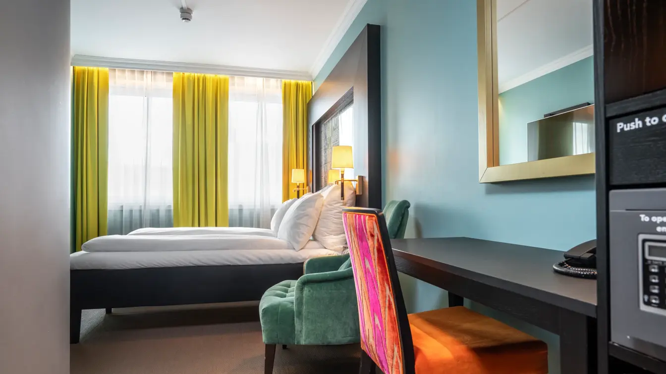 Dobbeltrom med twin senger, skrivebord med oransje stol, grønn lenestol, gule gardiner og turkis vegg.
