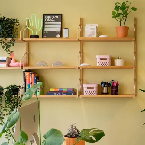 Detaljer fra vegg på kontor med gule vegger, grønne planter og vegghengt hylle med hjemmekoselige ting.