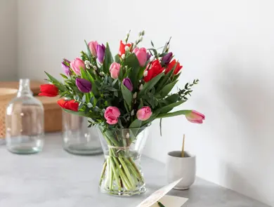 Stor bukett tulipaner står i vase på et bord, med glass og kurver bak. Noen gåsunger blant alle tulipanene i ulike farger. 