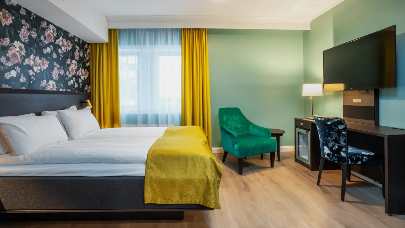 Turkise vegger, mørkt skrivebord med veggehengt TV, og mønstrete stol. Grønn lenestol. Vindu med gul gardin. Dobbeltseng med gult sengeteppe. I dobbeltrom på Thon Hotel Høyers i Skien.